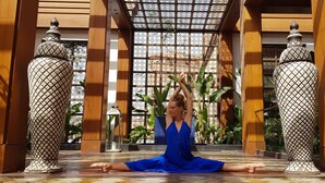 Una persona con un vestido azul realizando una postura de yoga en un amplio espacio interior con pilares de madera y suelo estampado.