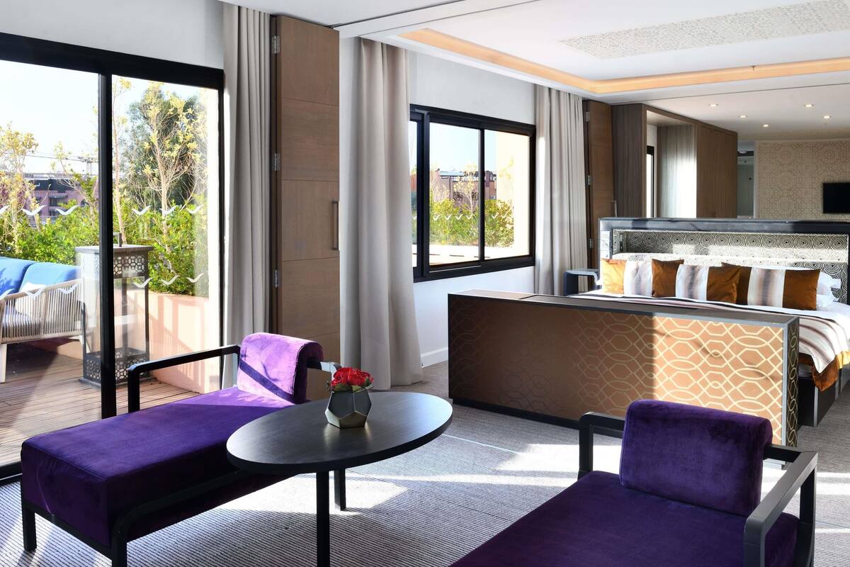 Ein Hotelzimmer mit Balkon, einem Bett mit gemusterten Textilien, einem kleinen Tisch mit Blumenvase und violetten Sitzgelegenheiten.