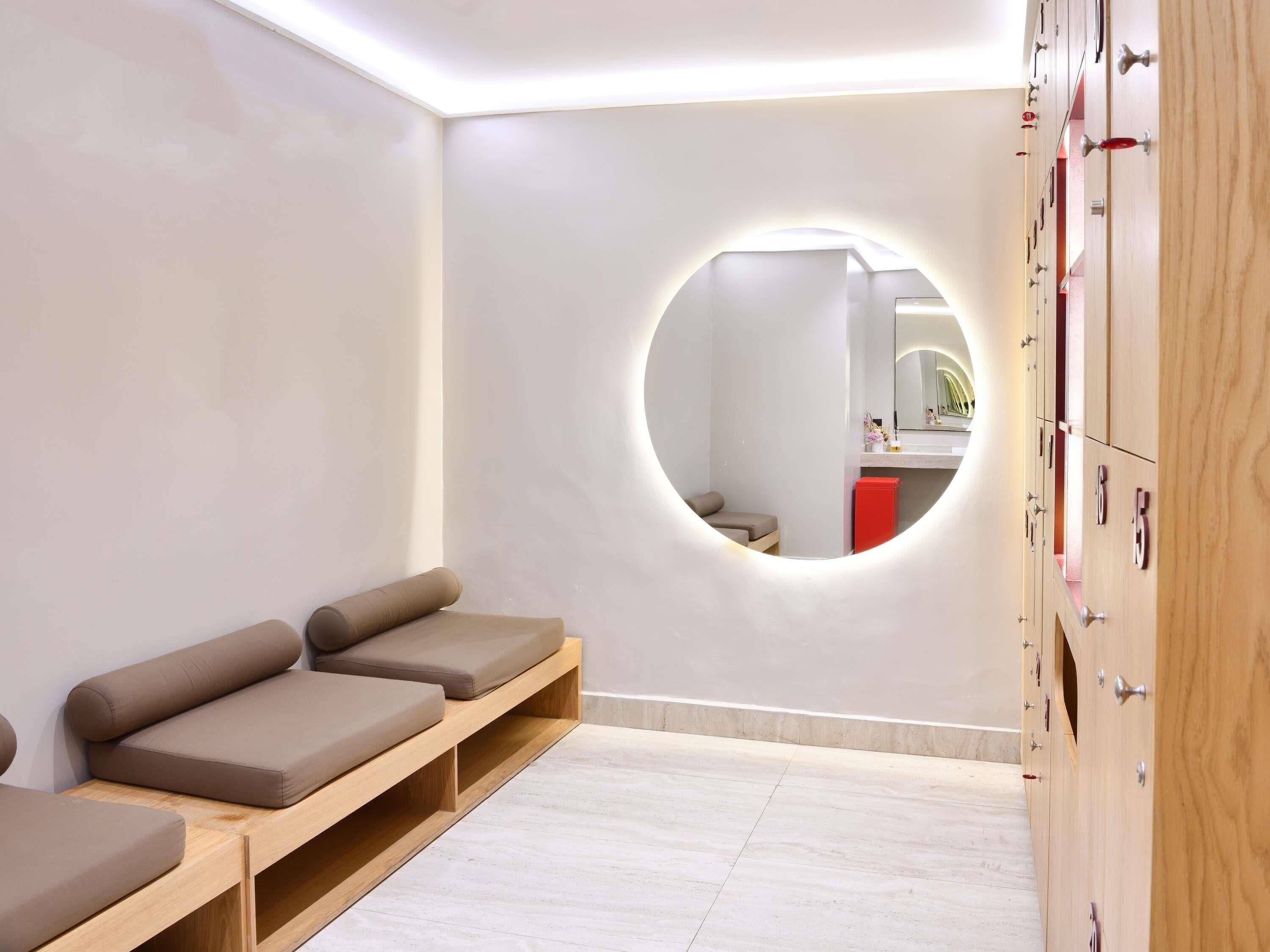 A modern spa room with a circular mirror, wooden benches, and a wooden cabinet with drawers.