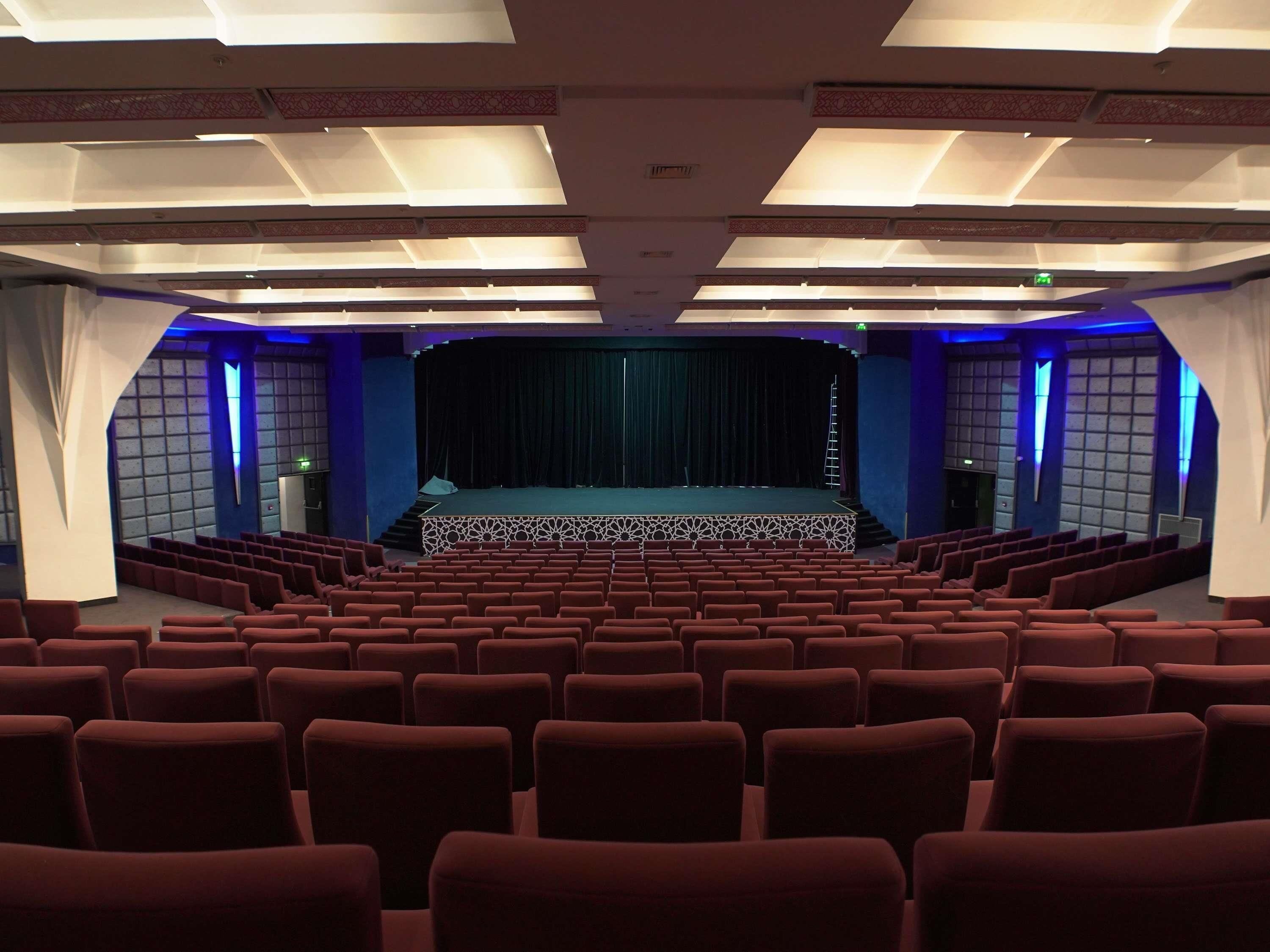 An indoor auditorium with rows of red seats, a stage with a curtain, and patterned ceiling lights.