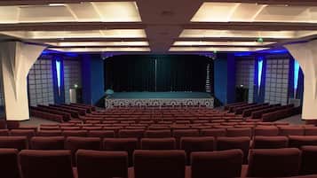 An indoor auditorium with rows of red seats, a stage with a curtain, and patterned ceiling lights.