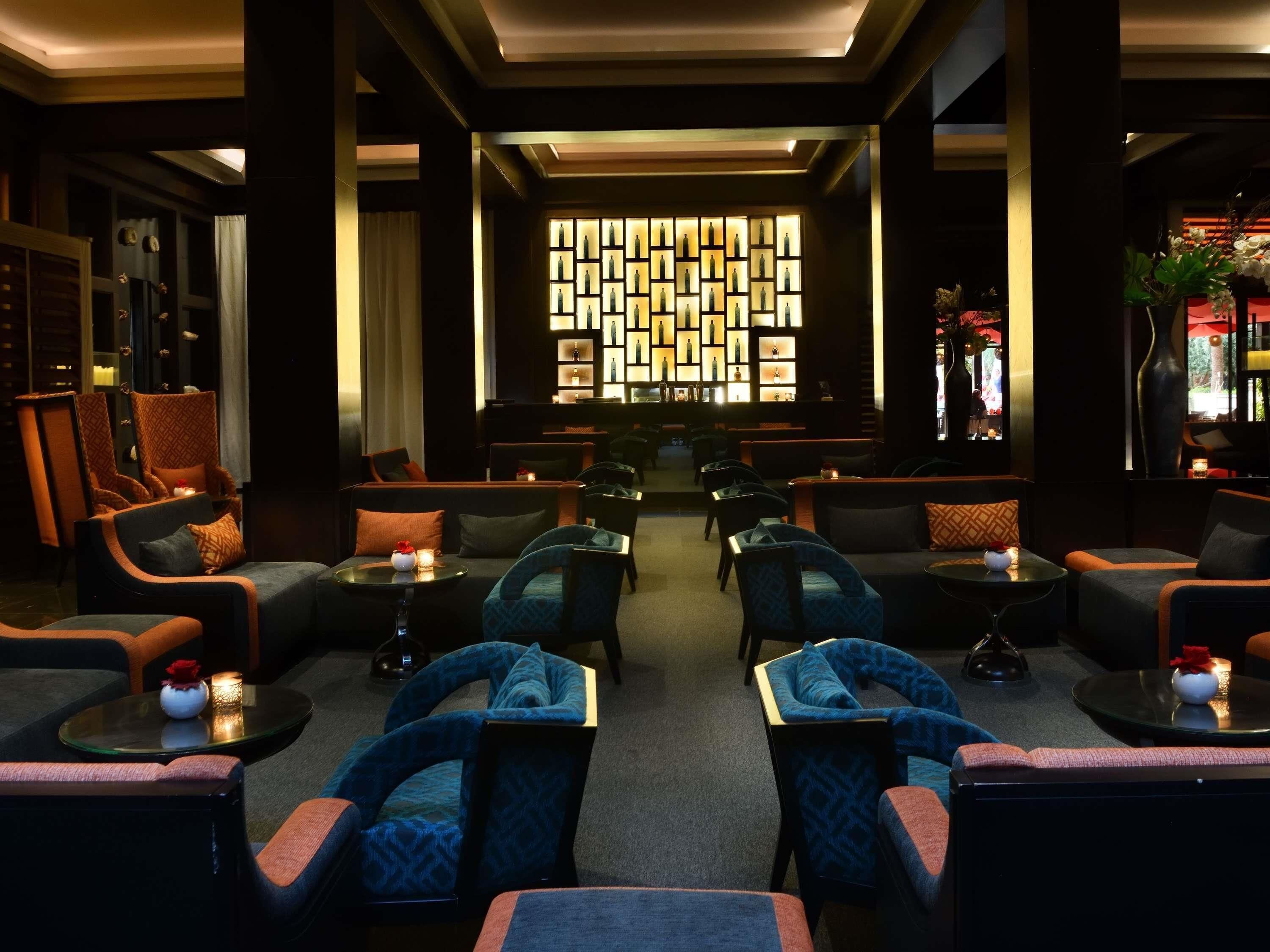 A dimly lit lounge area with multiple seating options including sofas and armchairs, a patterned wall feature, and a bar area in the background.