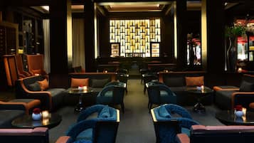A dimly lit lounge area with multiple seating options including sofas and armchairs, a patterned wall feature, and a bar area in the background.