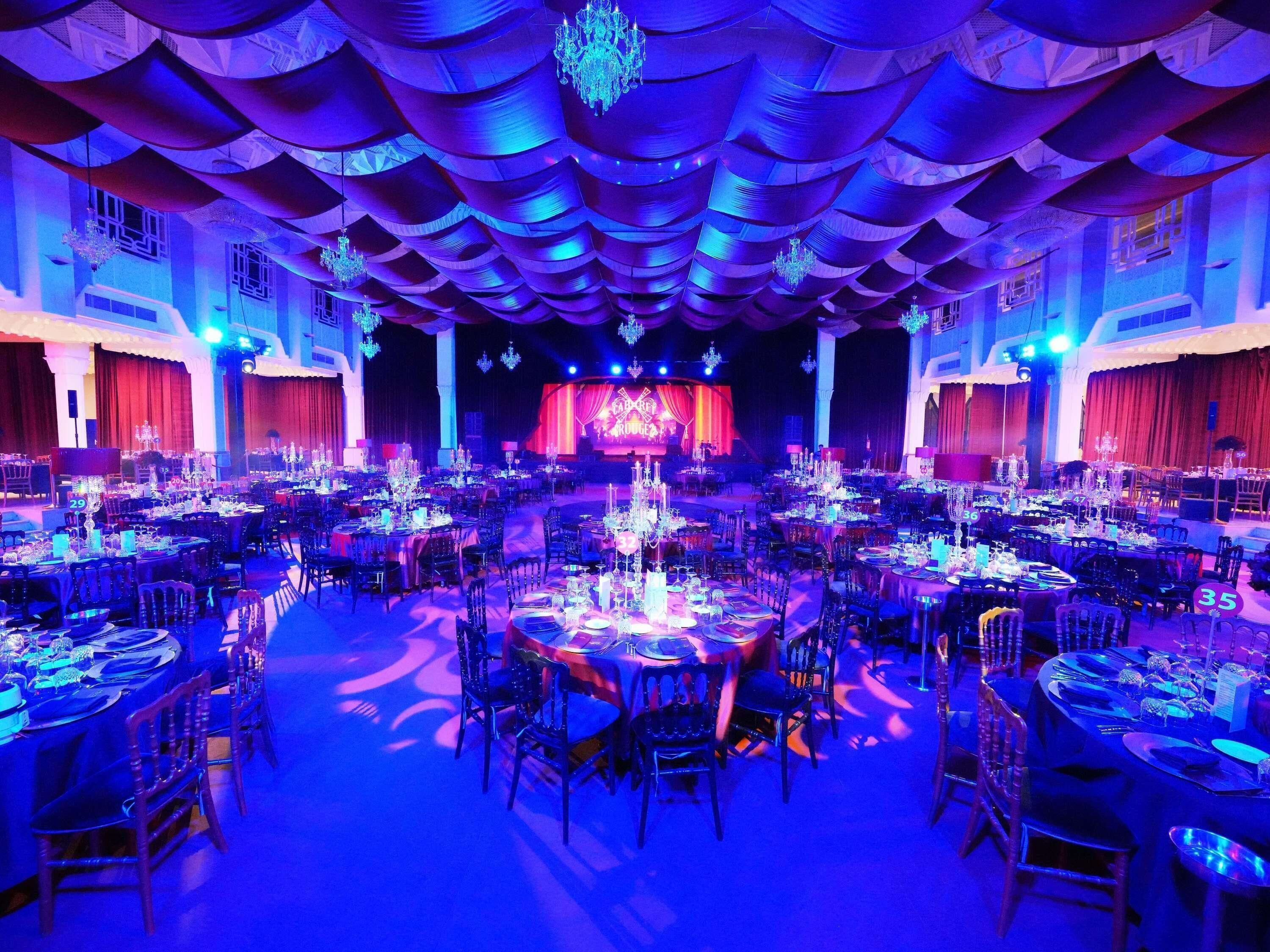 A large banquet hall with round tables, chandeliers, and a stage.