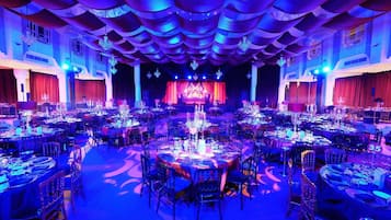 A large banquet hall with round tables, chandeliers, and a stage.