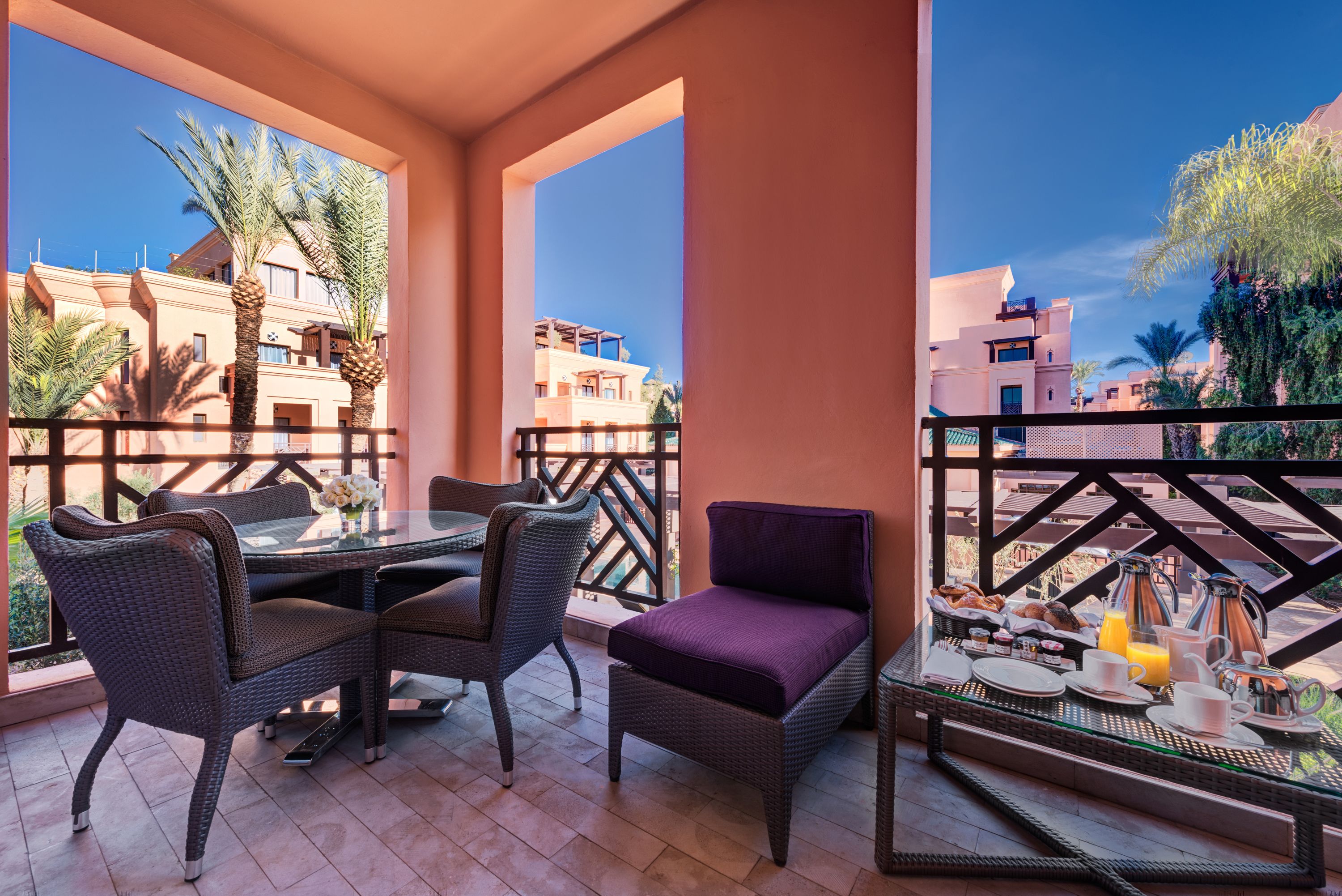A balcony with wicker furniture, a dining table set for breakfast, and a view of buildings and palm trees.