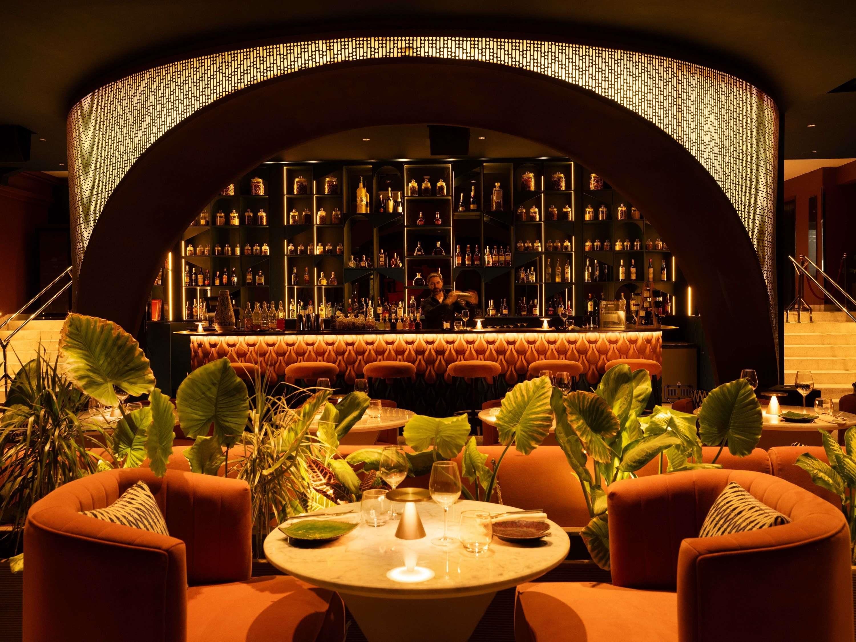 Zona de bar con una pared curva de estanterías llenas de botellas, un área de asientos con mesas redondas y sillas naranjas, y plantas verdes grandes.