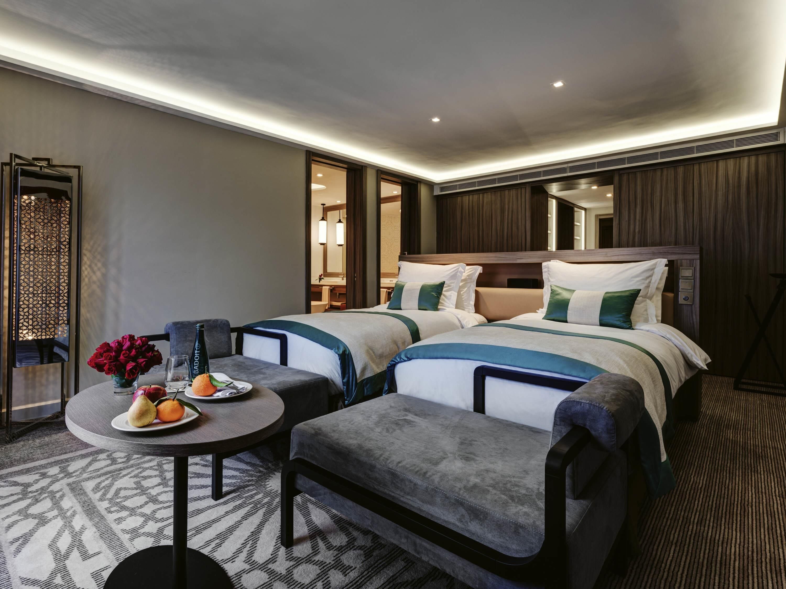 A hotel room with two beds, a small table with fruit, a chair, and a decorative mirror.