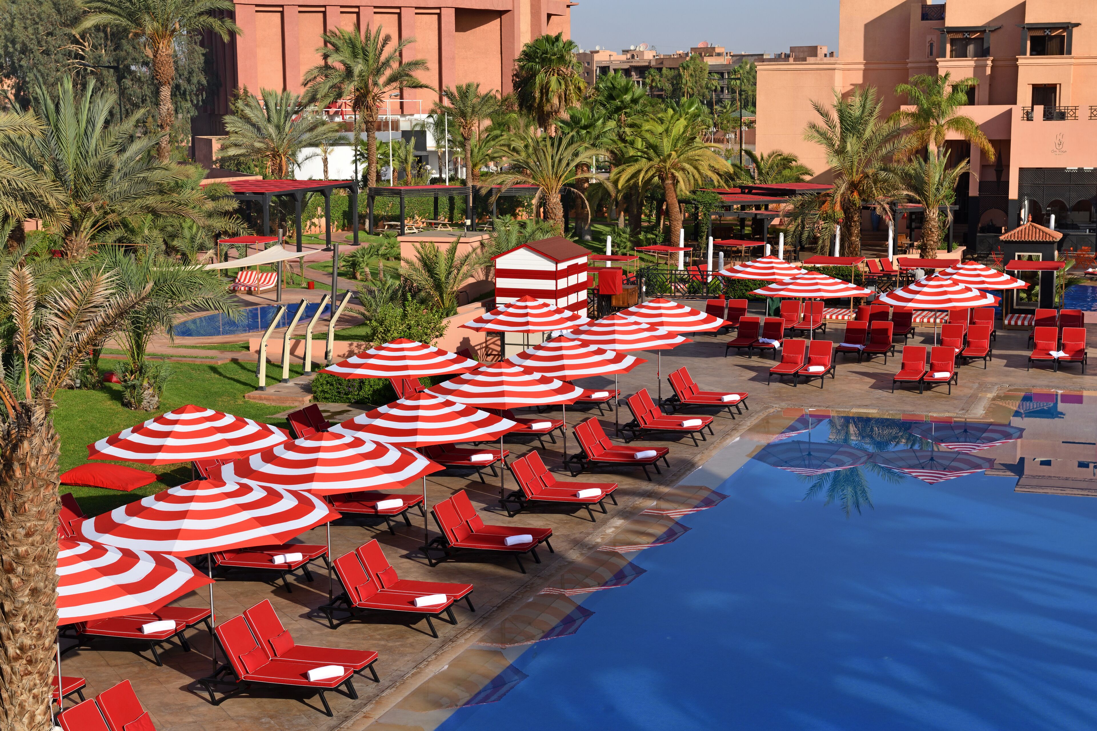 A poolside area with red lounge chairs and striped umbrellas, surrounded by palm trees and a building in the background.