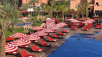 A poolside area with red lounge chairs and striped umbrellas, surrounded by palm trees and a building in the background.