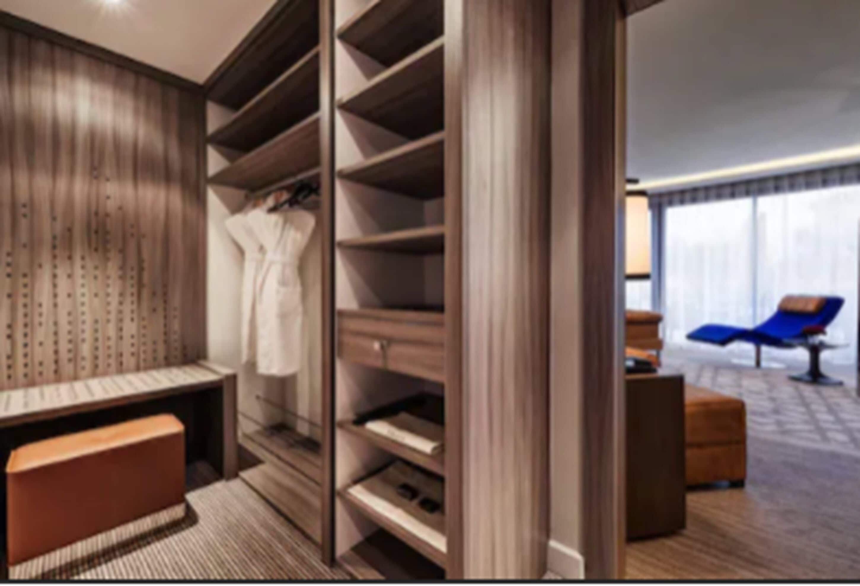 A modern hotel room with a large walk-in closet, a seating area with a bench, and a view of the city through large windows.