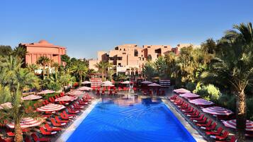 A pool with red lounge chairs and umbrellas, surrounded by palm trees and buildings.