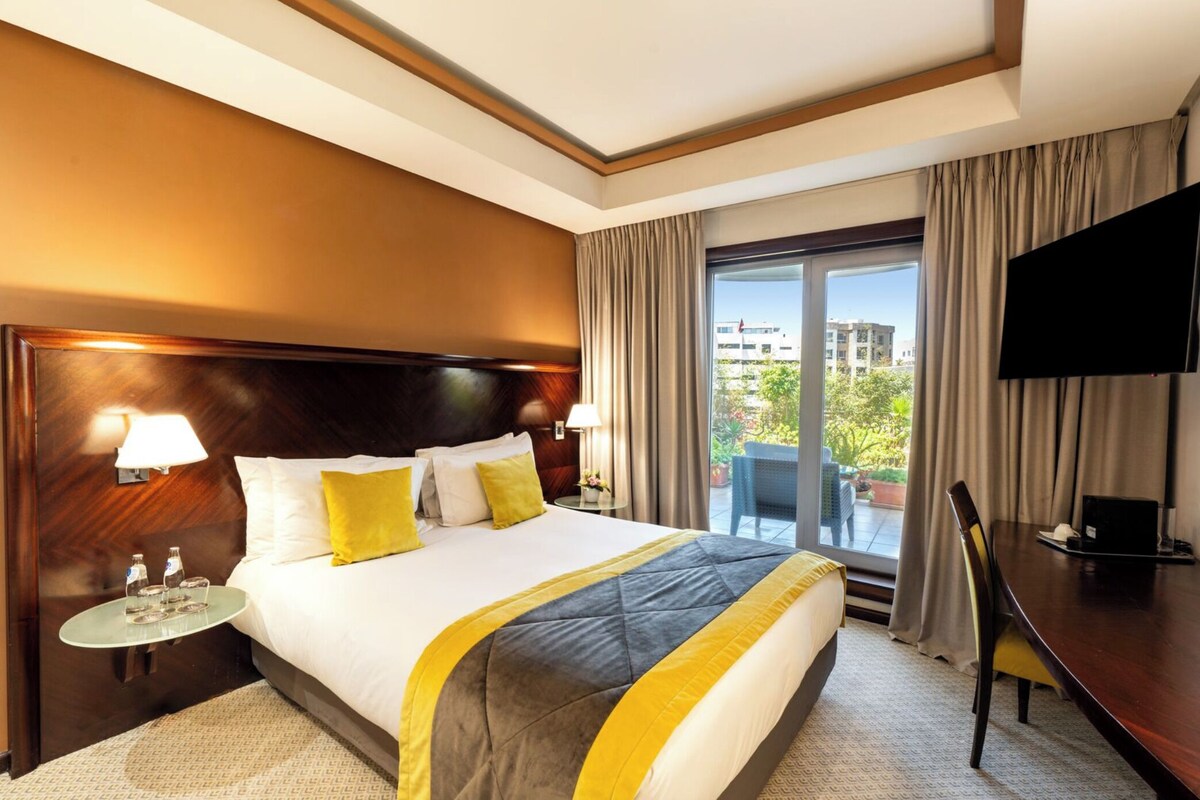 A hotel room with a large bed, a TV, a desk, and a balcony with a view of buildings.