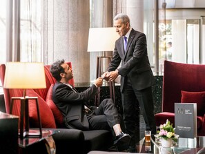 Ein Mann in einem Anzug schüttelt die Hand eines anderen Mannes, der in einem Hotel mit rotem Sofa und Glastisch mit einem Getränk sitzt. Ein Schild zeigt 'ACCORD LIVE LIMITLESS'.