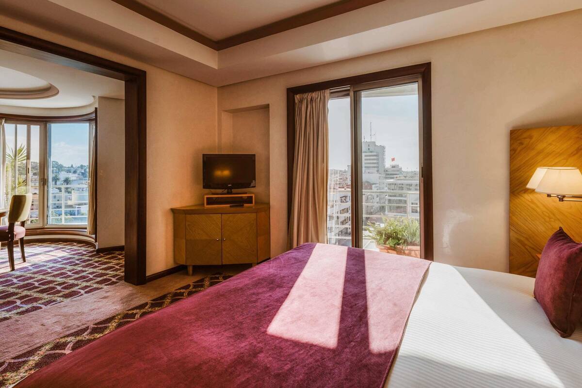 A hotel room with a bed, a television on a wooden cabinet, a lamp, and a view of the city through large windows.