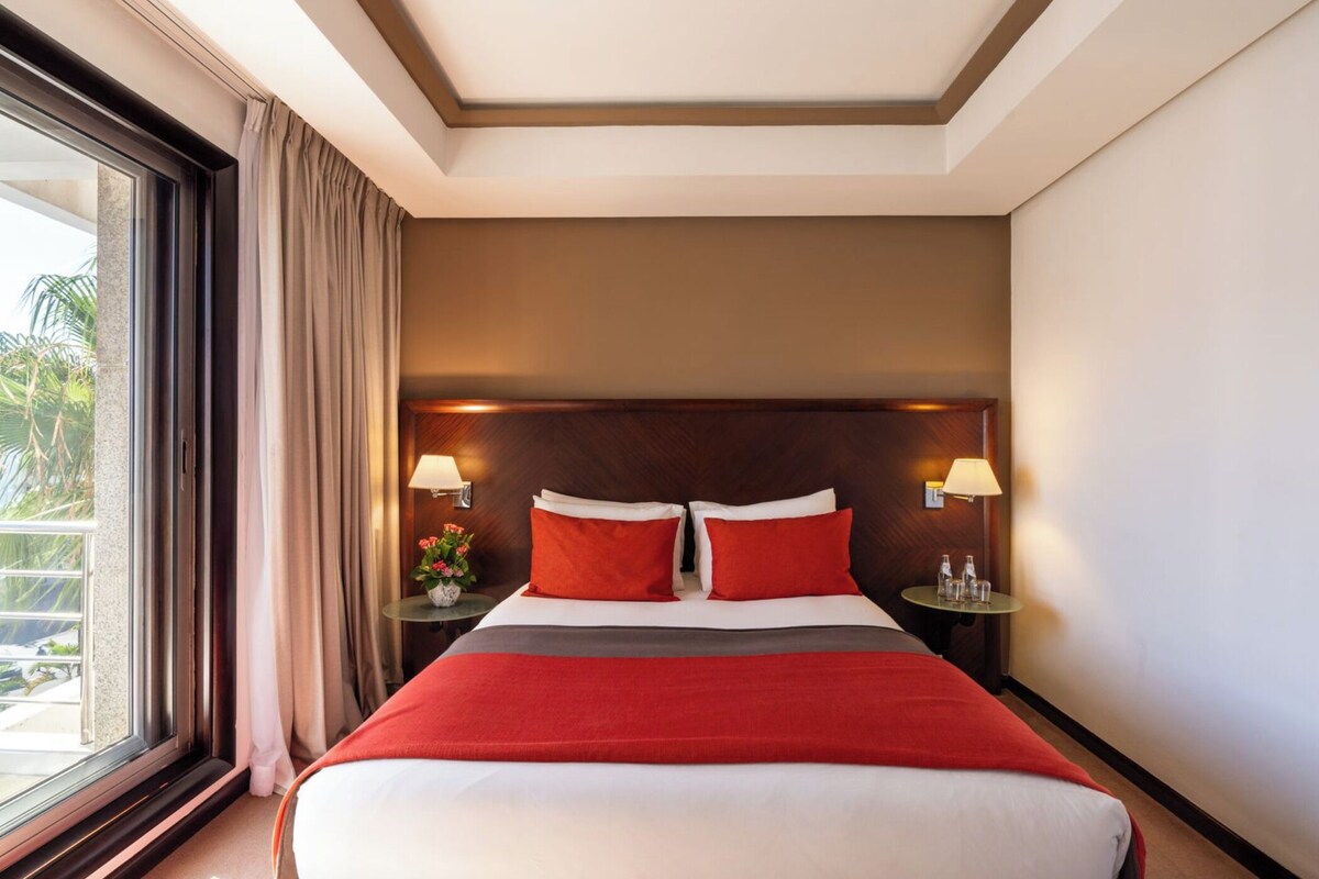 A hotel room with a large bed, two bedside lamps, a window with curtains, and a view of palm trees.