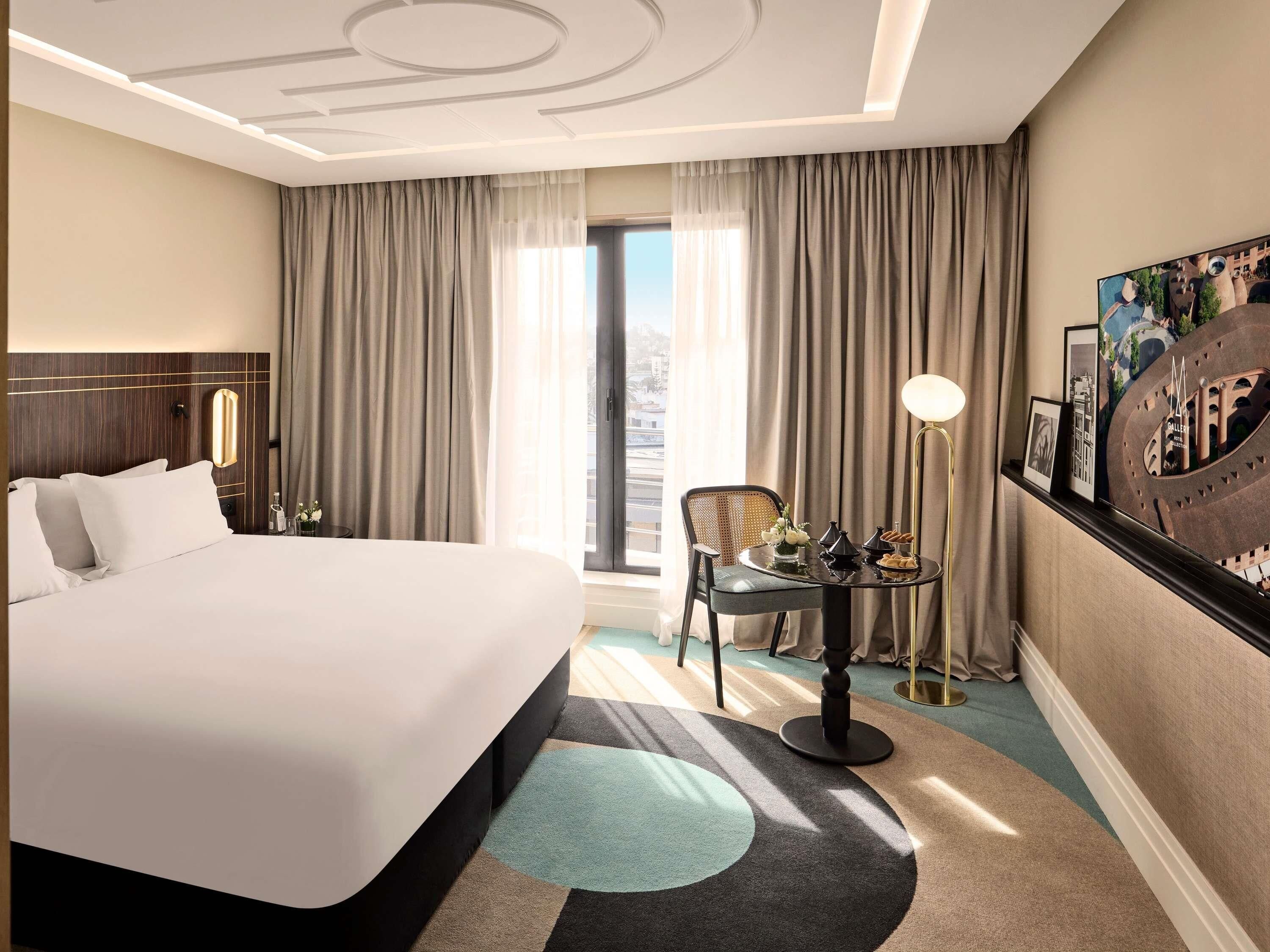 A hotel room with a large bed, a desk with a lamp, a chair, and a view of the city through the window.