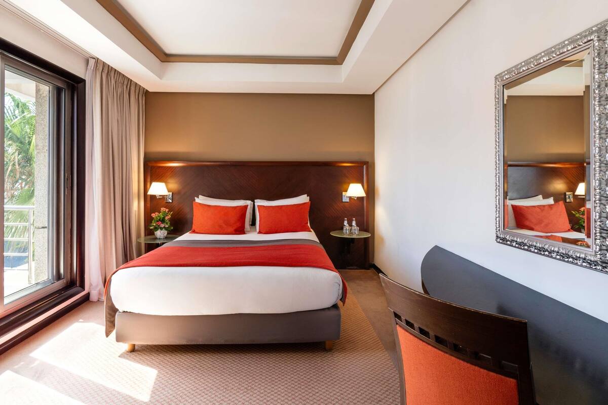 A hotel room with a large bed, two bedside lamps, a round table, a mirror, and a view of greenery outside.