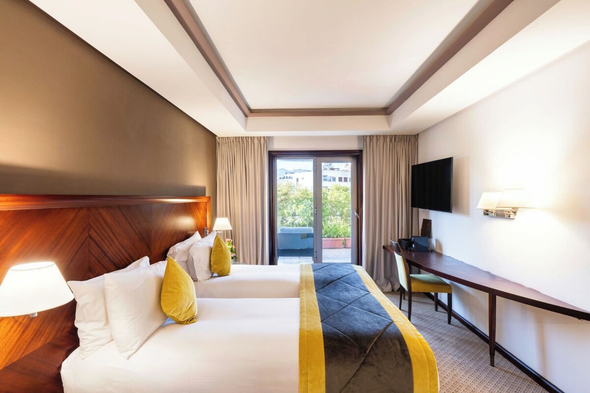 A hotel room with two beds, a desk, a TV, and a balcony with curtains.