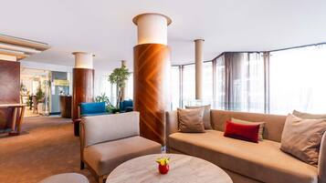 Un moderno hall de hotel con una gran columna de madera, un sofá color crema y una mesa redonda de mármol con un florero.