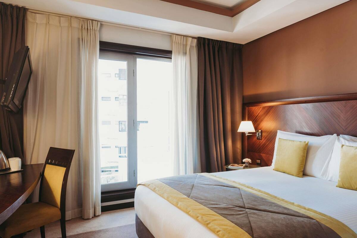 A hotel room with a bed, a desk, a chair, a lamp, and a view through the window.