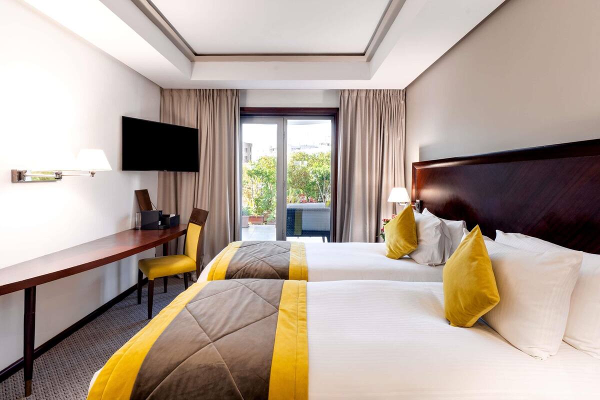 A hotel room with two beds, a desk, a chair, a TV, and a view of the outdoors.