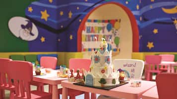 A children's play area with a birthday cake, chairs, and a bookshelf in the background.