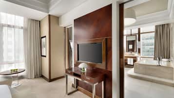 Habitación de hotel con un televisor de pantalla plana grande montado en un panel de madera, una pequeña mesa redonda con un jarrón de flores, un sofá y un ventanal con cortinas.