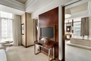 Habitación de hotel con un televisor de pantalla plana grande montado en un panel de madera, una pequeña mesa redonda con un jarrón de flores, un sofá y un ventanal con cortinas.