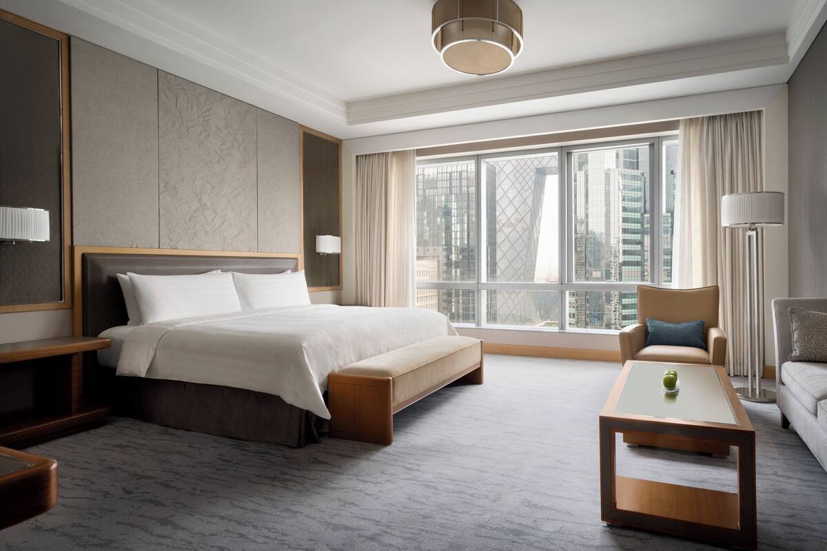 A modern hotel room with a large bed, a sitting area with a coffee table, and a view of the city through large windows.