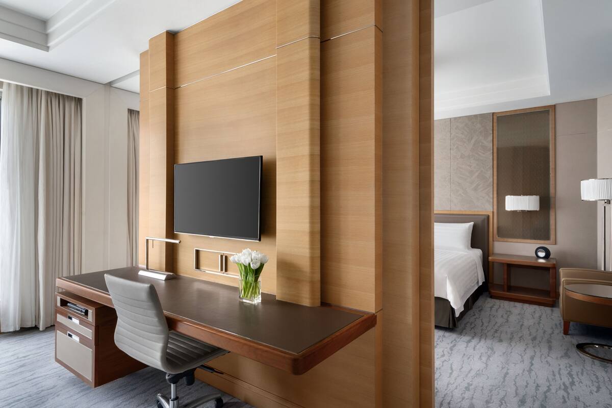 A modern hotel room with a wooden desk, a flat-screen TV, a bed, a bedside table with a lamp, and a large mirror.