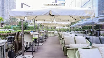Een terras met tafels en stoelen onder parasols, met een uitzicht op gebouwen op de achtergrond.