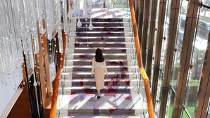 Una mujer baja una escalera con piso estampado, rodeada de barandas de vidrio y lámparas colgantes.