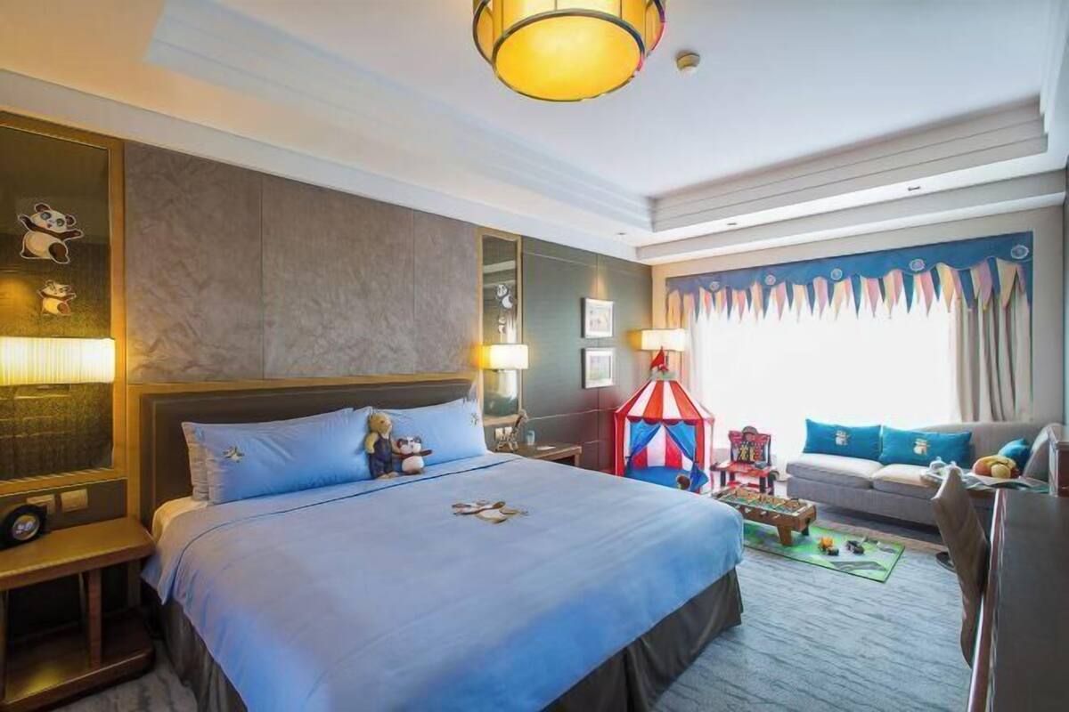 A hotel room with a large bed, bedside lamps, a small table, a colorful play tent, and a sofa with cushions.
