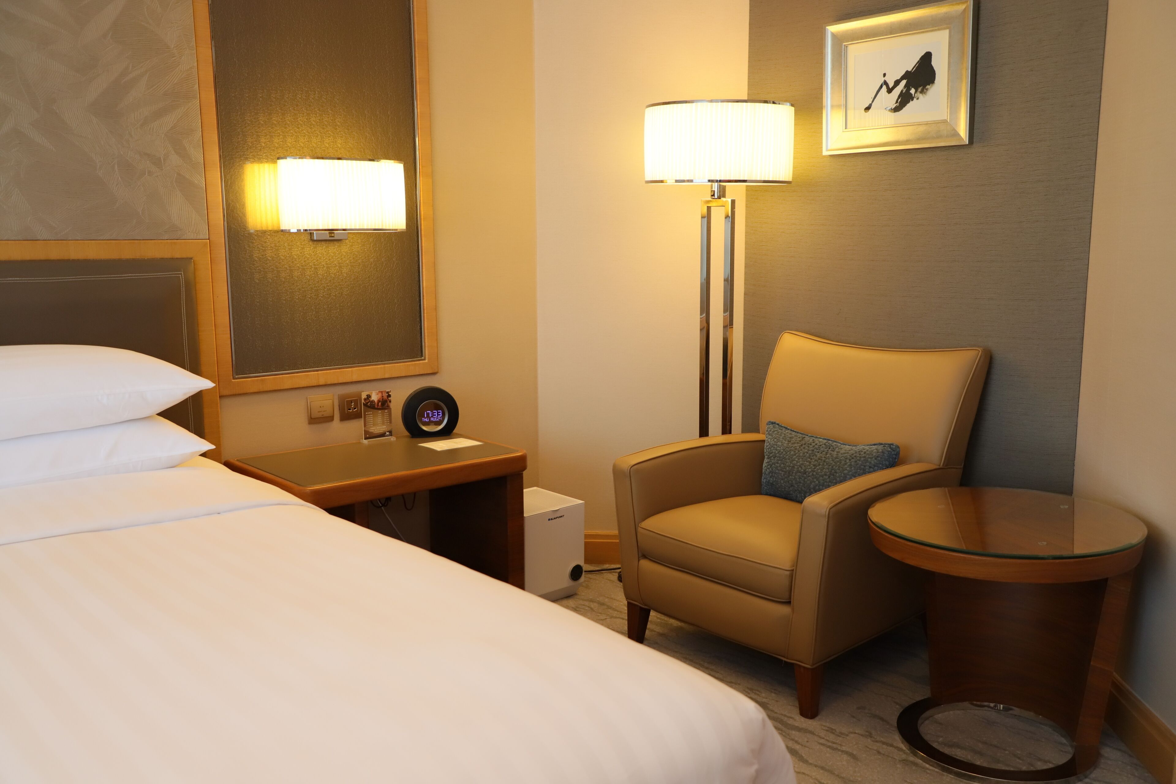 A hotel room with a bed, a desk, a chair, a lamp, a picture on the wall, and a small trash bin.