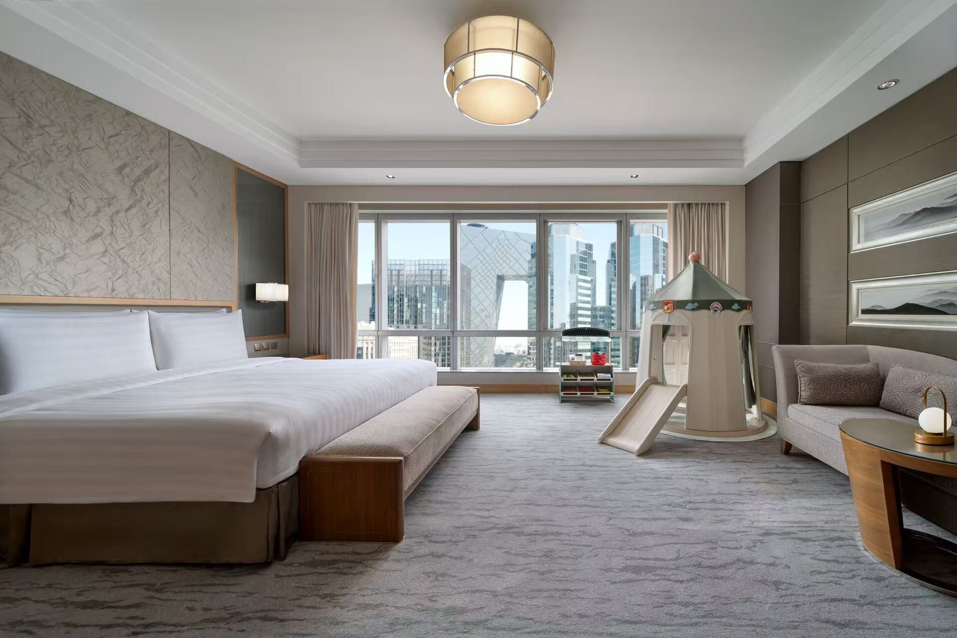 A hotel room with a large bed, a sofa, a small table, a lamp, and a view of the cityscape through large windows.