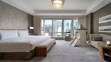 A hotel room with a large bed, a sofa, a small table, a lamp, and a view of the cityscape through large windows.