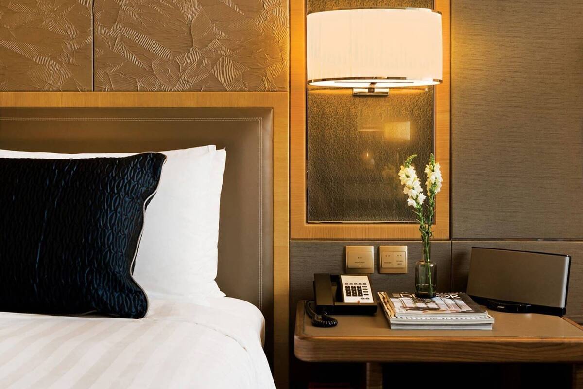 Habitación de hotel con cama, mesita de noche con lámpara, un jarrón con flores y escritorio con teléfono y revista.