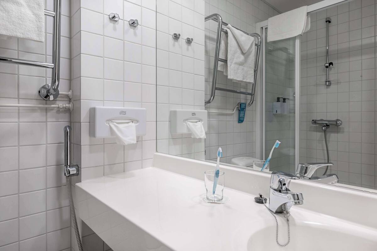 A modern bathroom with a white tiled wall, a glass shower enclosure, a white sink with a chrome faucet, and a glass soap dispenser.