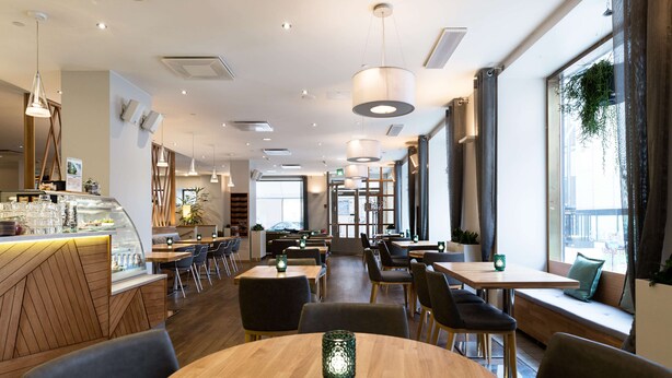 A modern restaurant interior with wooden tables, cushioned seating, pendant lighting, and large windows. - Scandic Plaza Turku (Turku)