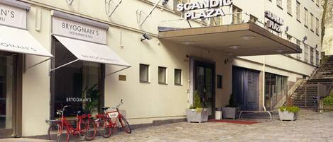 A building with a sign reading "Scandi Plaza" and "Bryggman's Restaurant & Deli". There are bicycles parked outside and potted plants on the sidewalk.