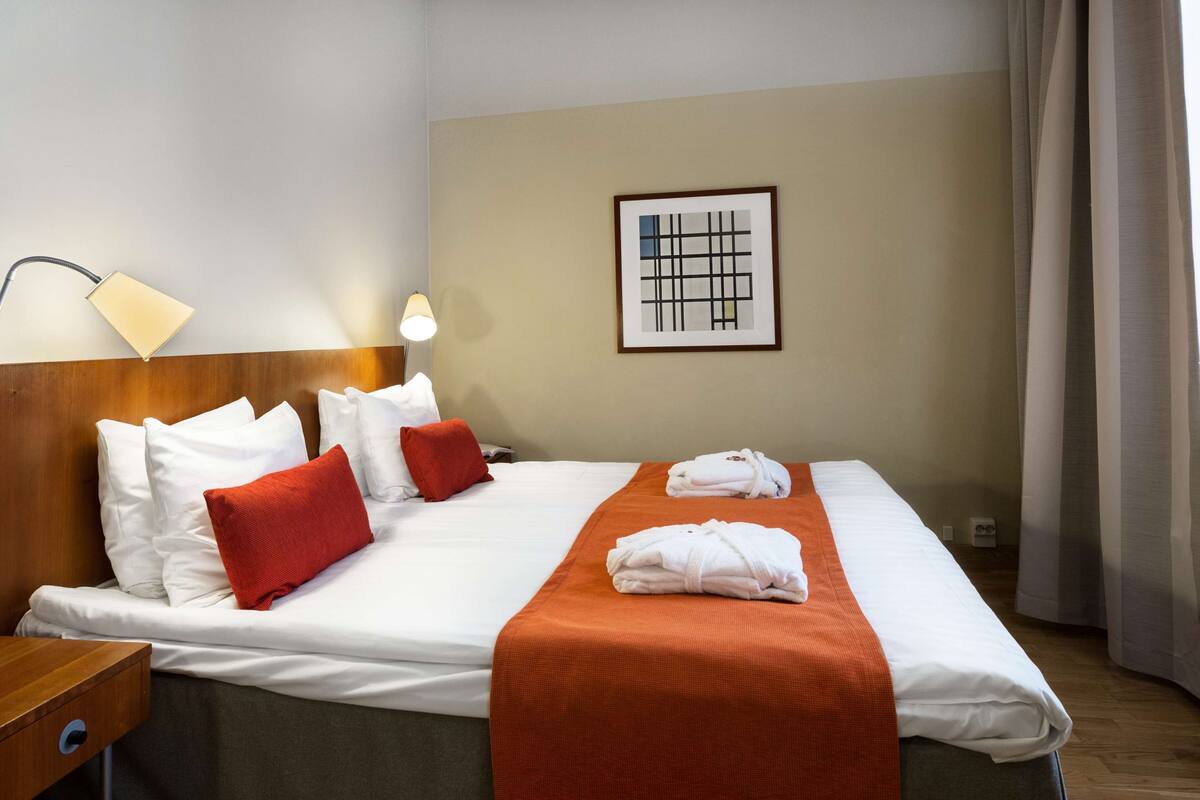 A neatly made bed with white and red pillows, an orange blanket, and two towels. There is a framed abstract artwork on the wall, a lamp on the nightstand, and a wooden headboard.