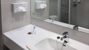 A modern bathroom with a white sink, a mirror, a glass shower enclosure, and two paper towel dispensers.