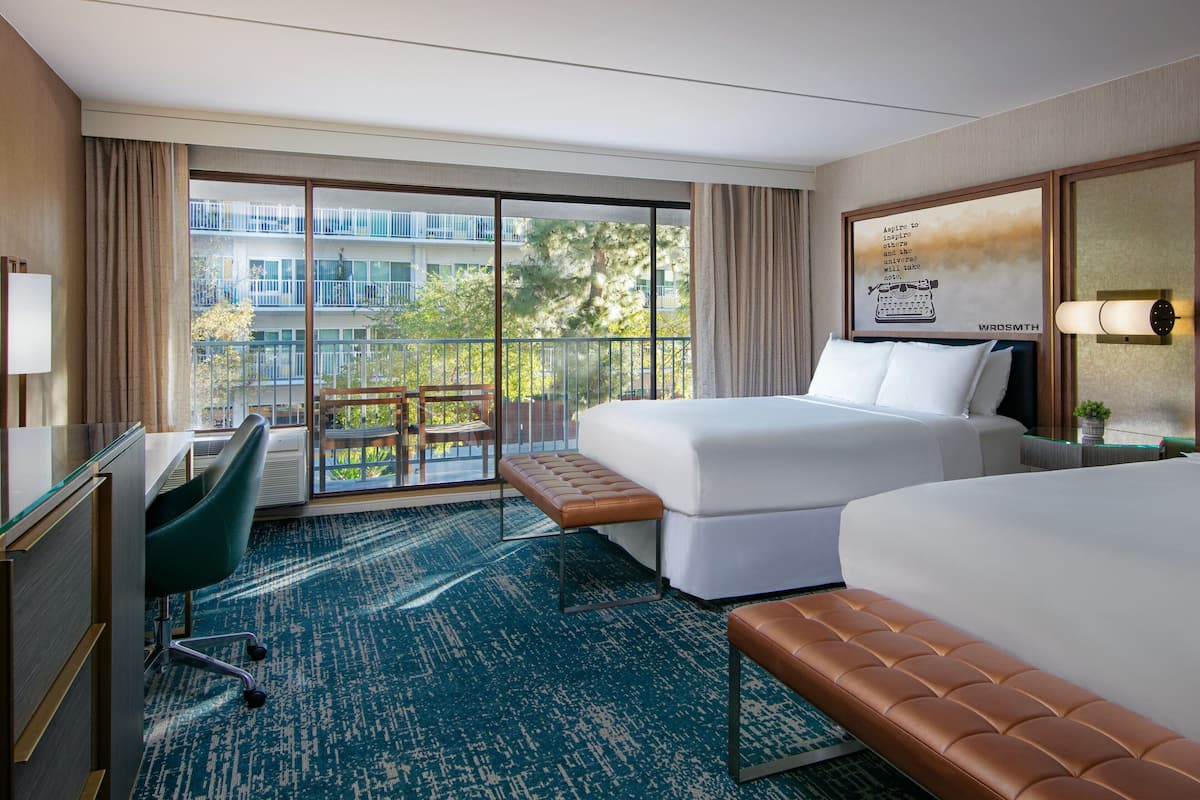 A hotel room with two beds, a desk, a chair, a bench, and a large window offering a view of a building and trees.