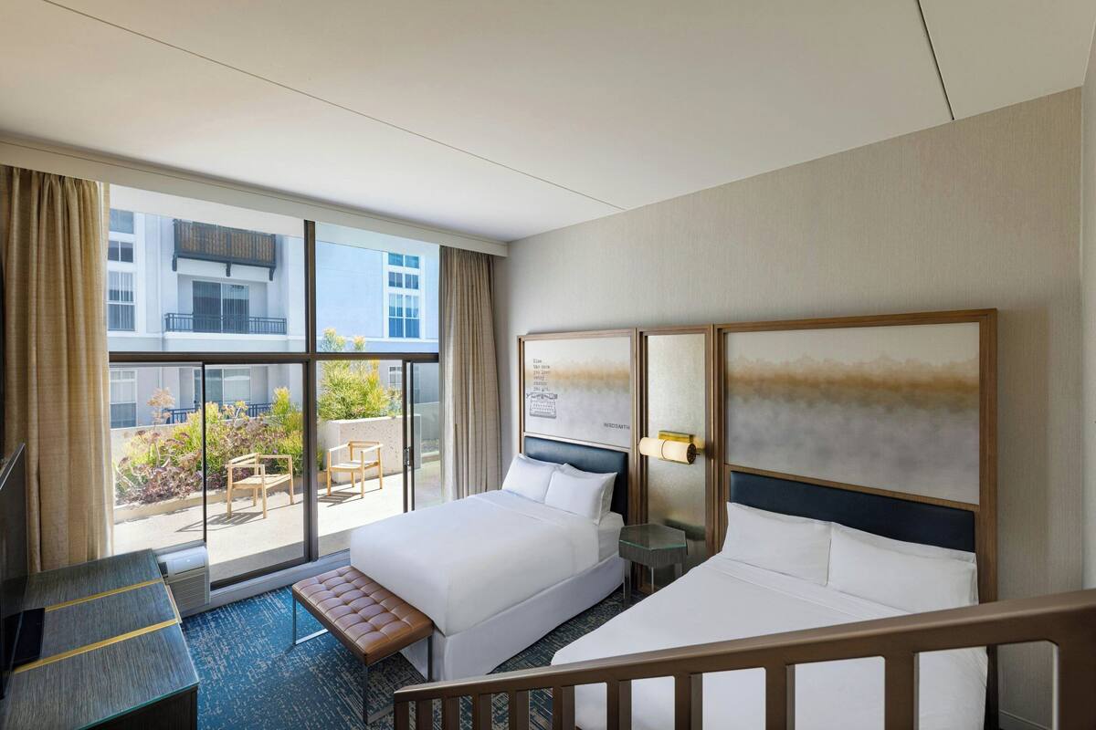 A hotel room with two beds, a desk, a bench, and a large window offering a view of the outside.
