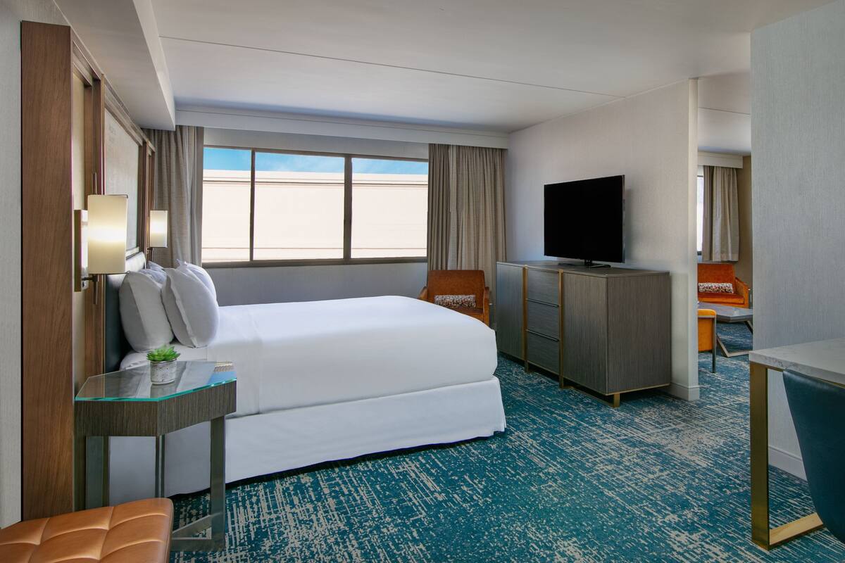 A hotel room with a large bed, a desk with a lamp, a television, and a view of the sky through the window.