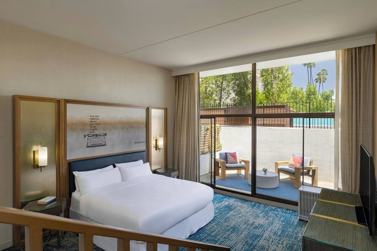 A hotel room with a large bed, a desk, a chair, and a TV. There is a balcony with a view of palm trees and a pool.