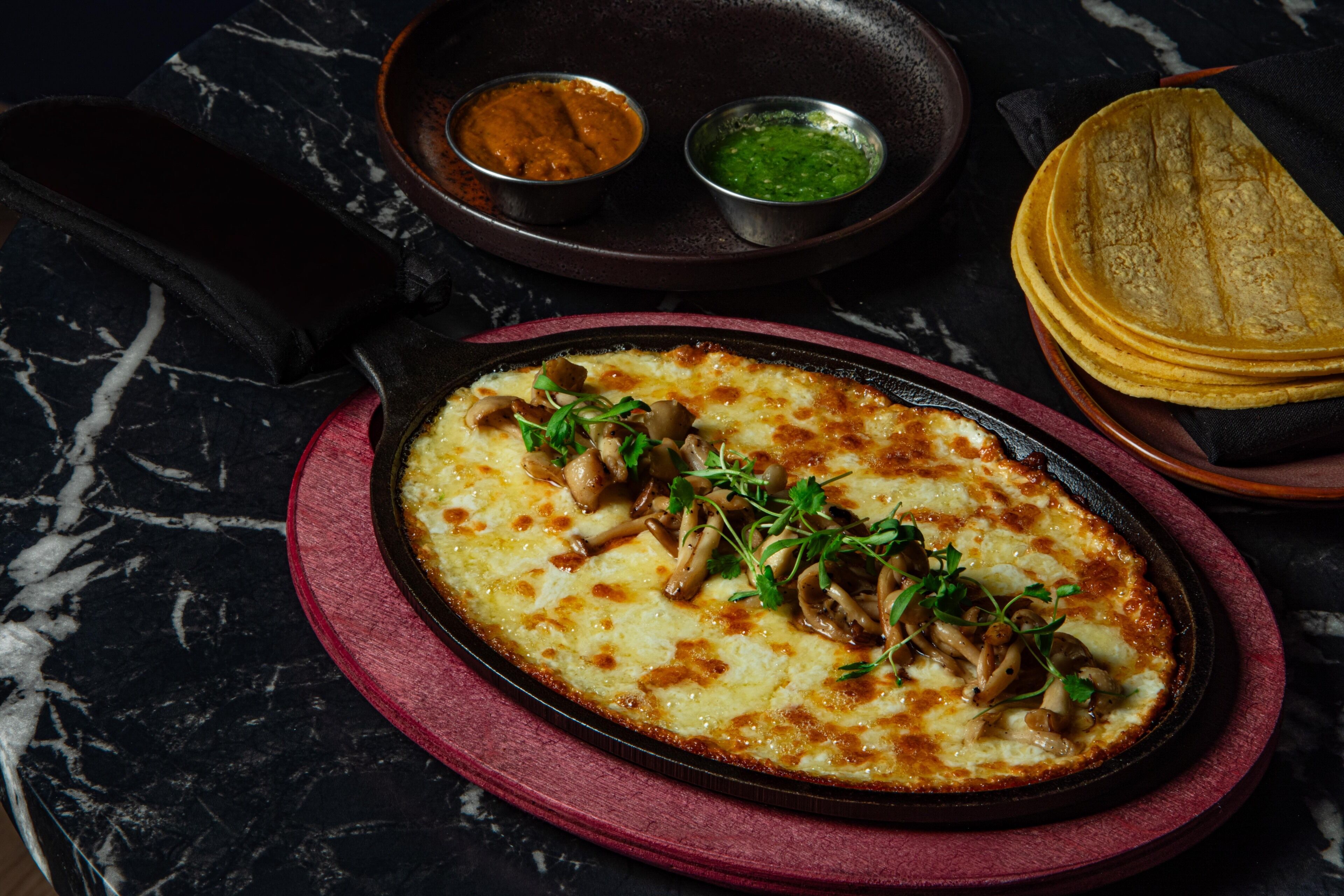 A dish with mushrooms and greens on a red plate, accompanied by two small bowls of sauce on a dark marble table.