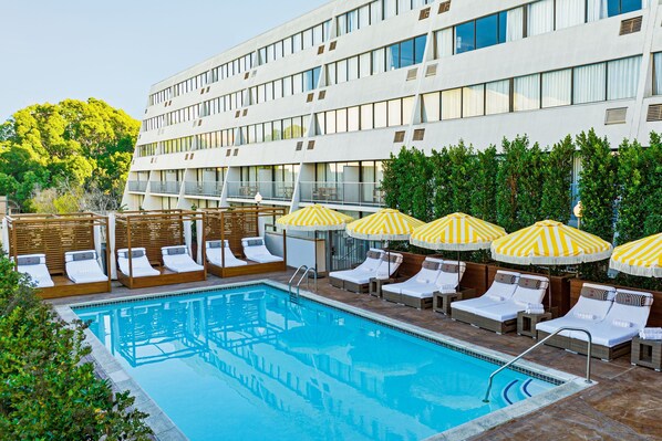 Una piscina con tumbonas y sombrillas, rodeada por un edificio moderno con amplias ventanas.