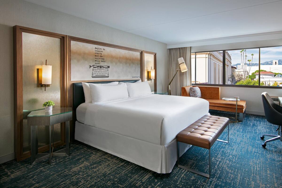 A hotel room with a large bed, a bench, a desk, and a view of the city.